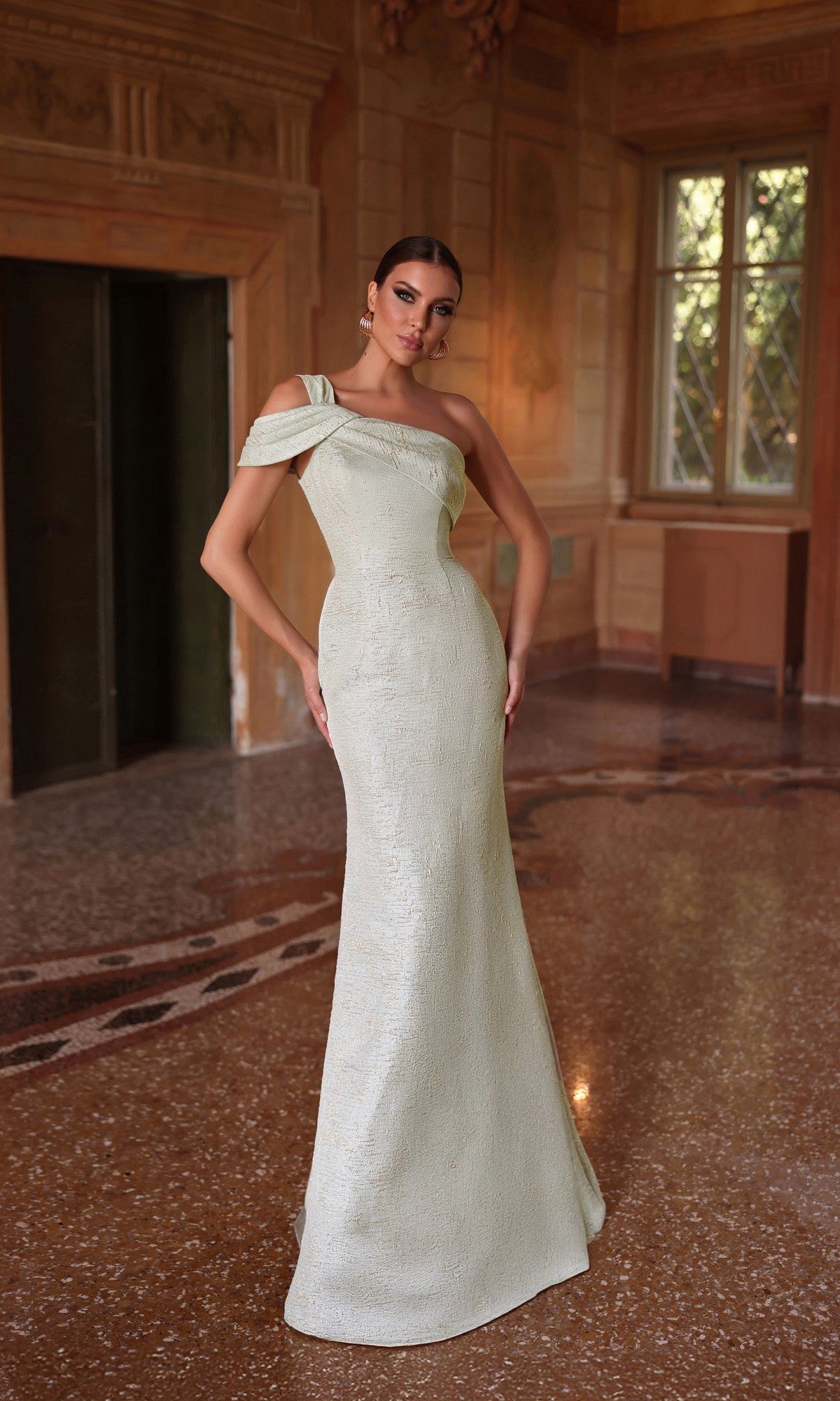 A woman in the Alyce 27749 Long Formal Dress, featuring a fitted sheath silhouette and cold-shoulder sleeves, stands in an elegant room with ornate walls and tall windows.